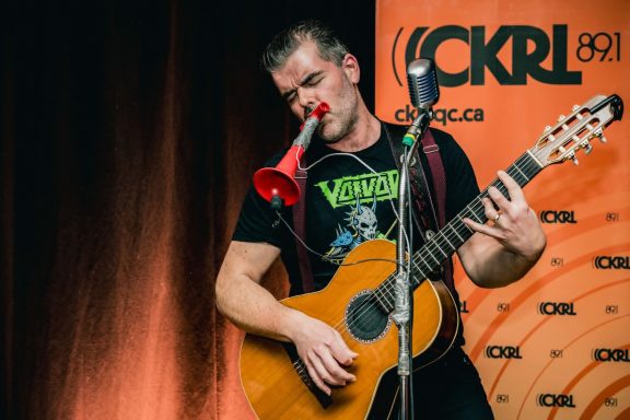 Pépé et sa guitare - Radiothon CKRL 89,1 Musician playing guitar and a kazoo, performing on stage with a microphone.