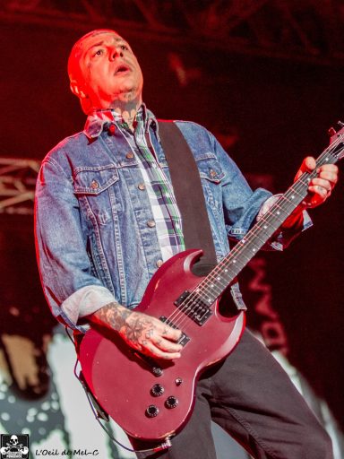 Musician playing electric guitar on stage, wearing a denim jacket and plaid shirt.