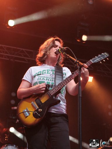 Musician with long hair singing into a microphone while playing guitar on stage.