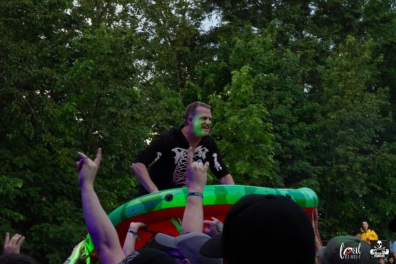 Person with face paint stands on a colorful float, surrounded by cheering crowd.