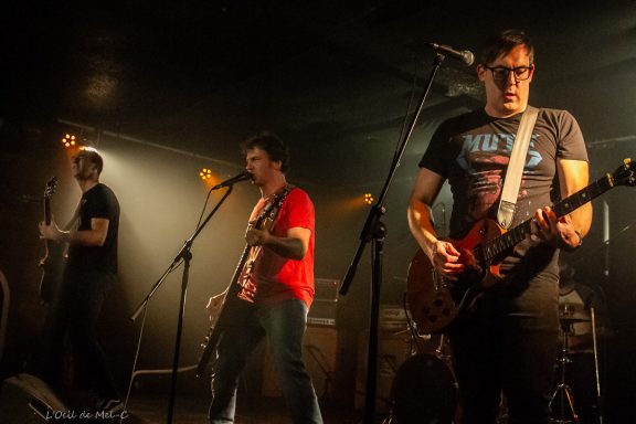 Three musicians performing on stage with guitars and microphones in a dimly lit venue.