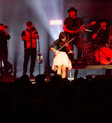A female violinist performs on stage with a band in a lively concert setting.