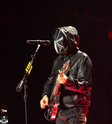 Person playing guitar on stage, wearing a hooded jacket and a mask with a geometric design.