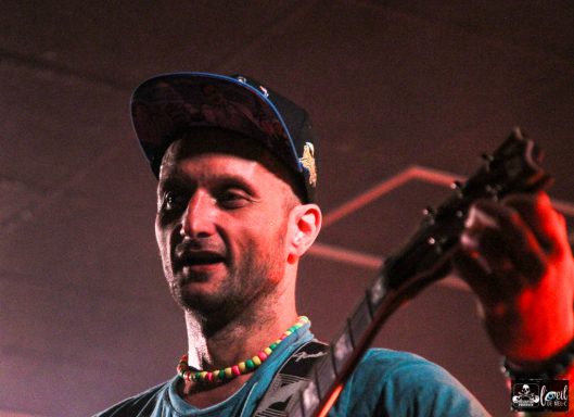 Straightline - L'Anti-Bar & Spectacles Musician performing on stage, wearing a cap and colorful accessories.