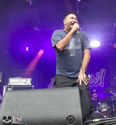 Man singing into a microphone on stage, with band members and colorful lights in the background.