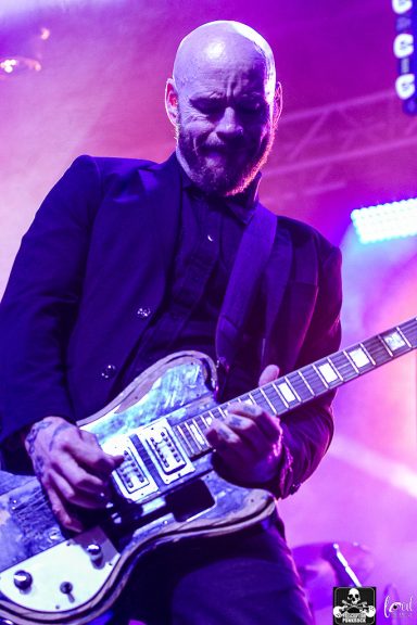 A musician performing on stage, playing an electric guitar with vibrant lights in the background.