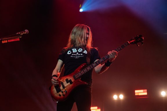 Musician playing bass guitar on stage with bright spotlights in the background.