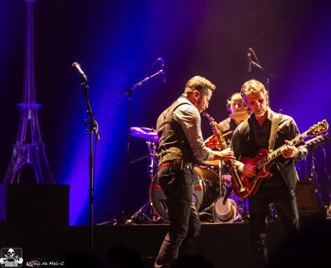 Musicians performing on stage with guitars and drums, illuminated by purple lights.