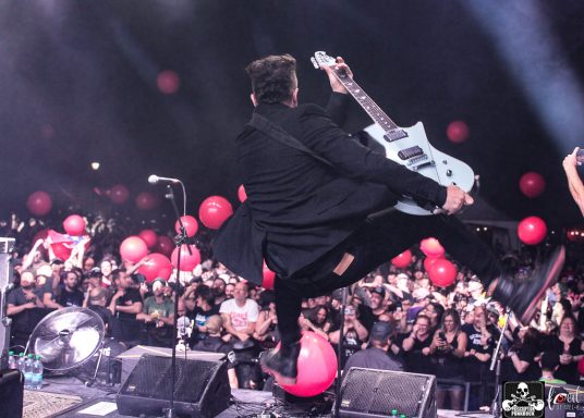 A guitarist leaps into the air on stage, with a crowd and red balloons in the background.