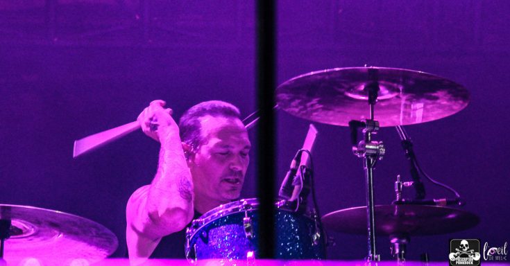 Drummer in a purple-lit setting, striking a cymbal with focused expression.