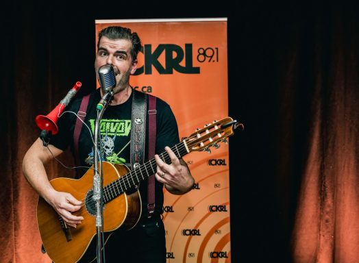 Pépé et sa guitare - Radiothon CKRL 89,1 Musician performing with an acoustic guitar and microphone in front of a radio station banner.