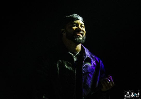 A man smiling with a dark background, wearing a leather jacket and a headband.