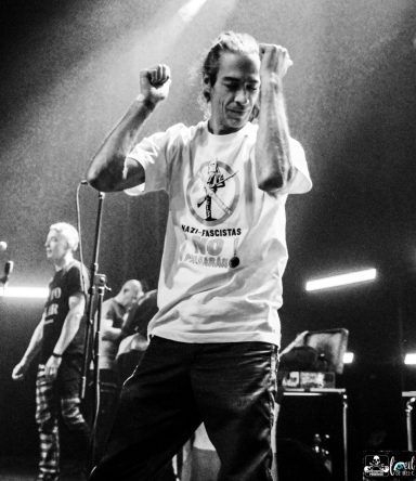 A performer energetic on stage, wearing a graphic t-shirt, with band members in the background.