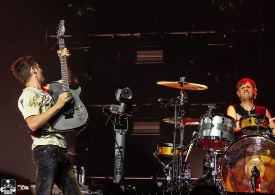 Musicians performing on stage; one plays guitar while the other drums.