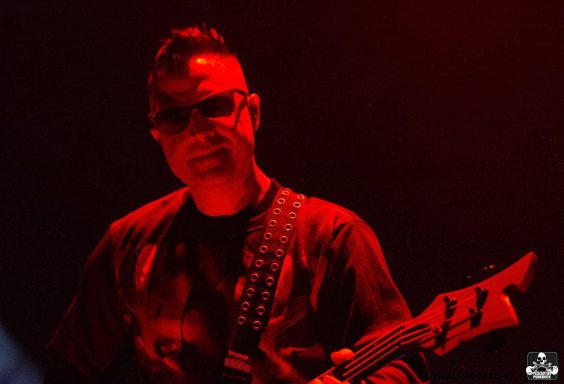 Musician in a red-lit setting, wearing sunglasses and playing an electric guitar.