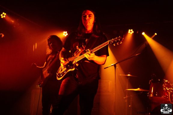 Musician playing guitar on stage with dramatic lighting and bandmates in the background.