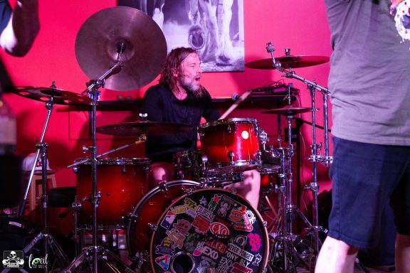 A drummer performs on stage, surrounded by colorful drum equipment and a vibrant backdrop.