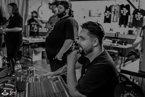 A sound engineer at a mixing console with people in the background, in a casual setting.