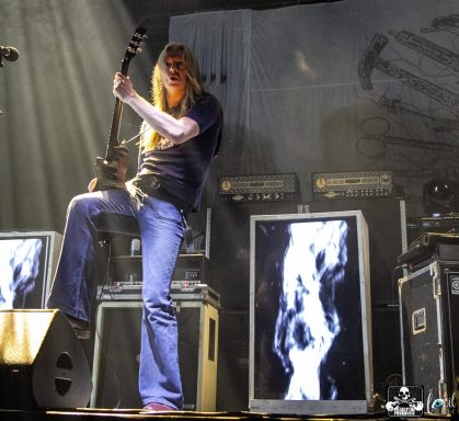 Musician playing guitar on stage with dramatic lighting and visual effects in the background.