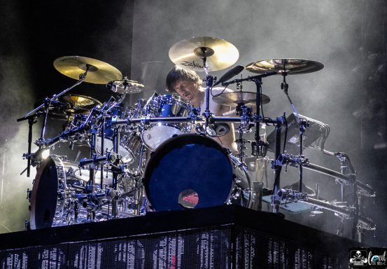 A drummer performing on stage with multiple drum kits and a smoky background.