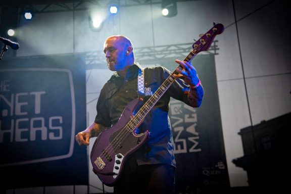 A musician playing a purple bass guitar on stage with blue lighting.