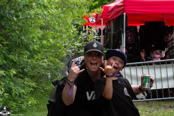 Two people making rock hand gestures, smiling, outdoors near a vendor booth.