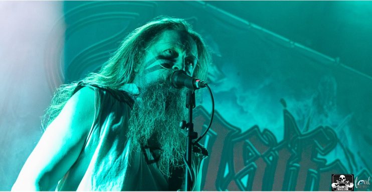 Musician with a long beard performing on stage, illuminated by green lighting.