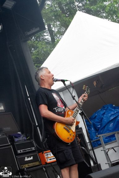 Man singing and playing guitar on stage with a large white tent in the background.