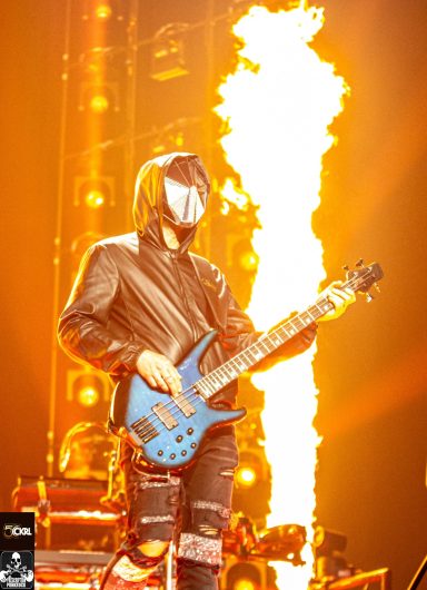 Musician in a dark outfit playing a blue guitar, with flames in the background.