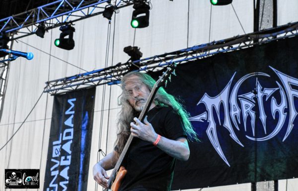 A guitarist performs on stage with a band logo in the background.