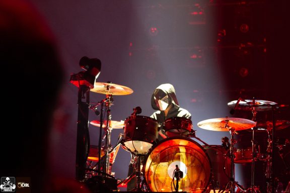 A drummer performing on stage with dramatic lighting and a silhouetted figure in the background.