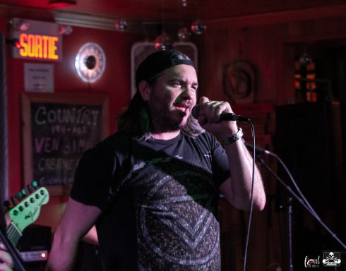 A performer singing into a microphone on stage, with band members visible in the background.