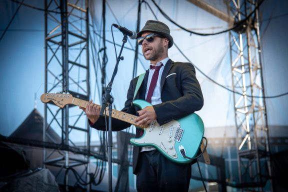 Man in a suit and fedora playing an electric guitar on stage.