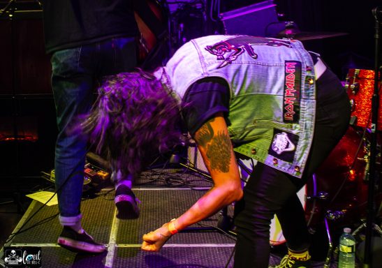 A musician bends down on stage, wearing a vest with patches and a tattoo visible on their arm.