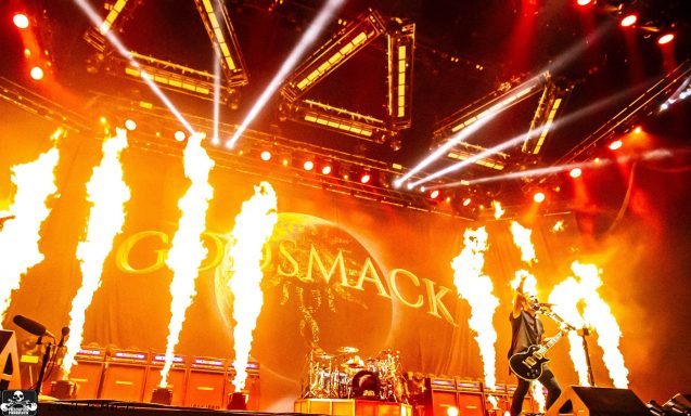 A guitarist performs on stage with bright lights and flames in a lively concert setting.