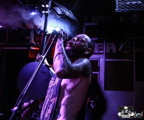 A tattooed man singing passionately on stage under purple lighting.