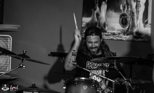 Musician playing drums, smiling, with long hair and a cap in a lively setting.