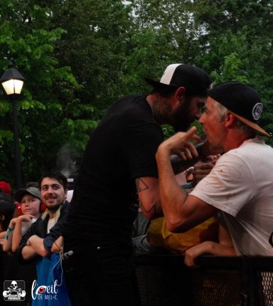 Two men engage in a lively conversation, surrounded by an excited crowd in a park.