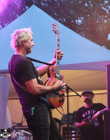 Musician playing guitar on stage with a bandmate in the background.