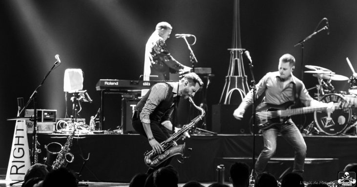 Live band performance with musicians playing saxophone and guitar, and a keyboardist in the background.