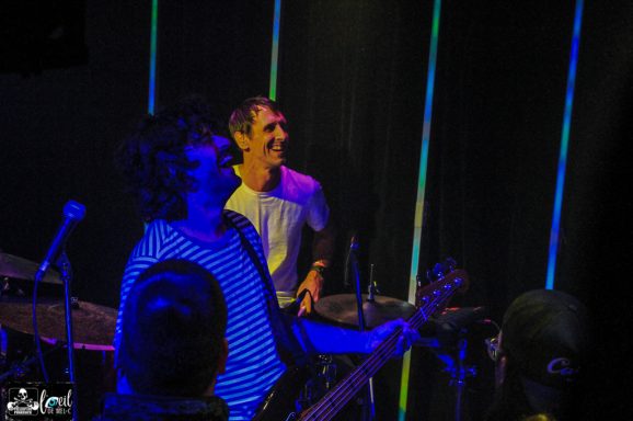 Two musicians perform on stage, with one playing guitar and the other on drums.