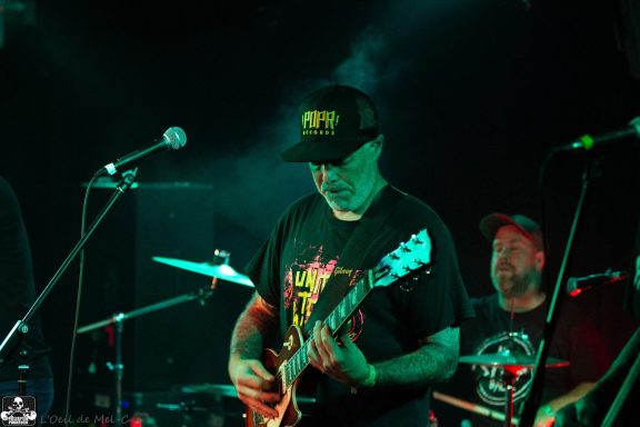 Musician playing guitar on stage with drums in the background. Colorful lighting.