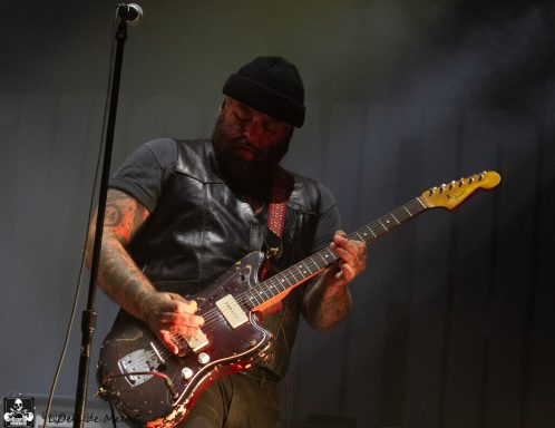 Man with a beard playing electric guitar on stage, wearing a black hat and vest.