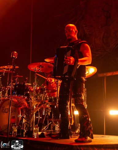 A musician playing the accordion on stage, surrounded by drums and dramatic lighting.