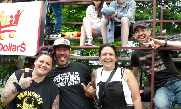 Four people smiling and posing outdoors, with a group sitting behind them.