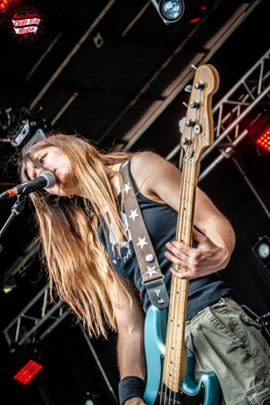 Musician playing an electric guitar on stage with long hair and a black tank top.