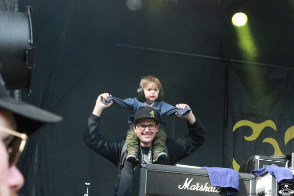 A person holds a child on their shoulders at a music event, both smiling and enjoying.