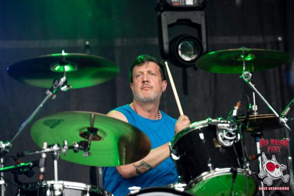 Drummer playing vigorously on stage with green lighting in the background.