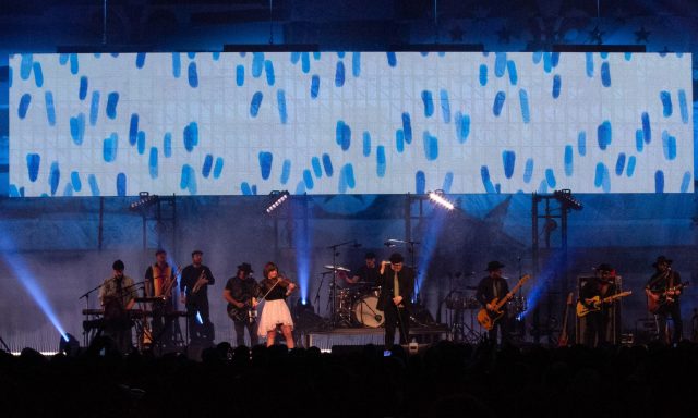 Musicians performing on stage with bright lights and a large screen in the background.