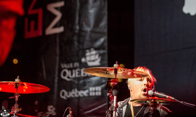 Drummer performing energetically on stage with a large banner behind.